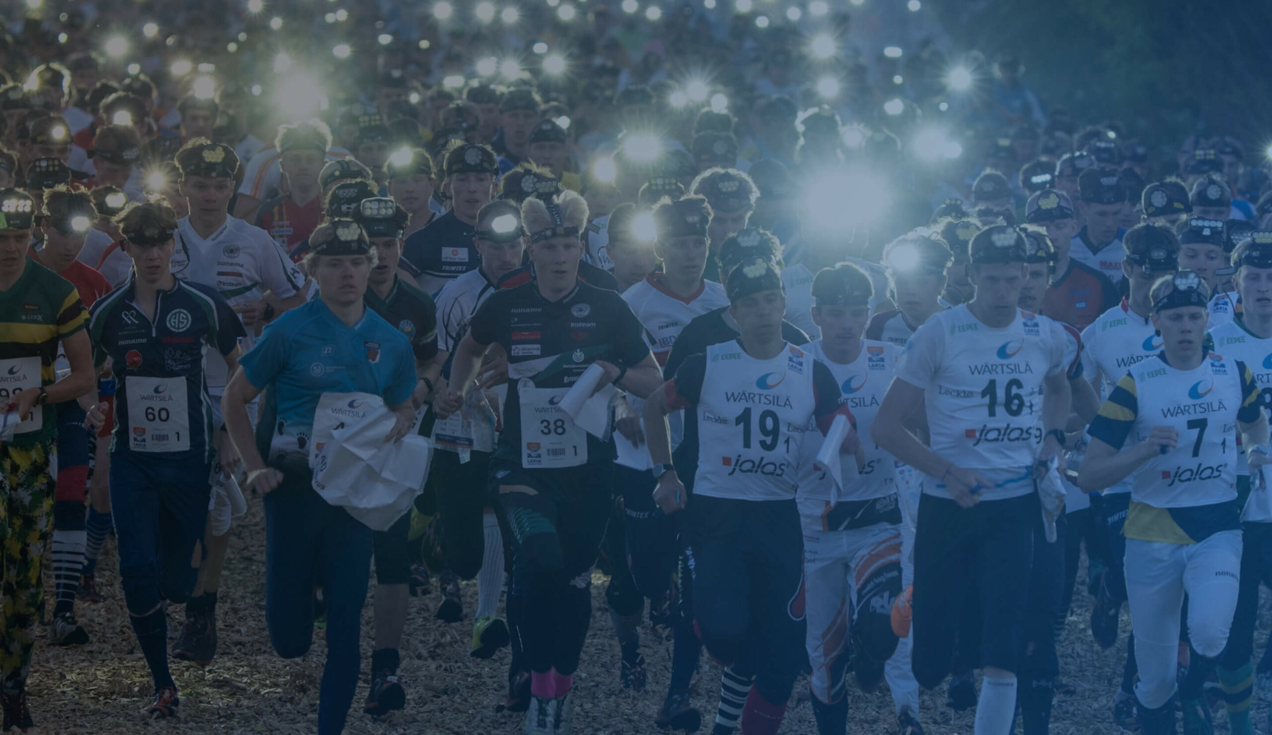 A large group of runners, many wearing headlamps and numbered shirts, participate in a nighttime orienteering race, holding maps as they move forward, with bright lights shining from their headlamps.