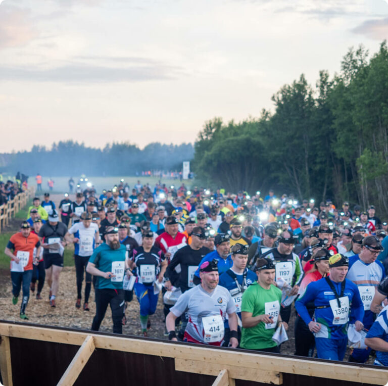 A large group of runners wearing headlamps and race bibs participate in an outdoor trail race at dusk, surrounded by trees and a wooden fence.