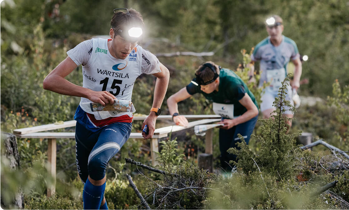 Three people wearing headlamps and numbered jerseys compete in an outdoor orienteering race through a wooded area, navigating with maps and checking control points.