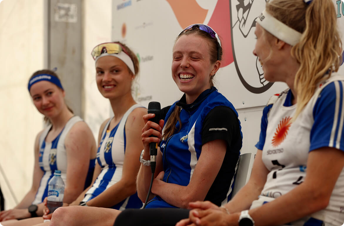 Four female athletes in blue and white sports uniforms sit and smile during what appears to be a panel or interview; one holds a microphone while others look on with relaxed, happy expressions.