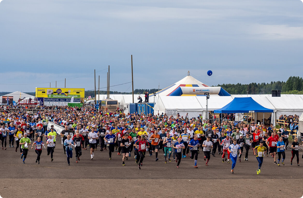 A large group of runners in colorful outfits start a race on a dirt track, with tents, banners, and spectators in the background under a cloudy sky.