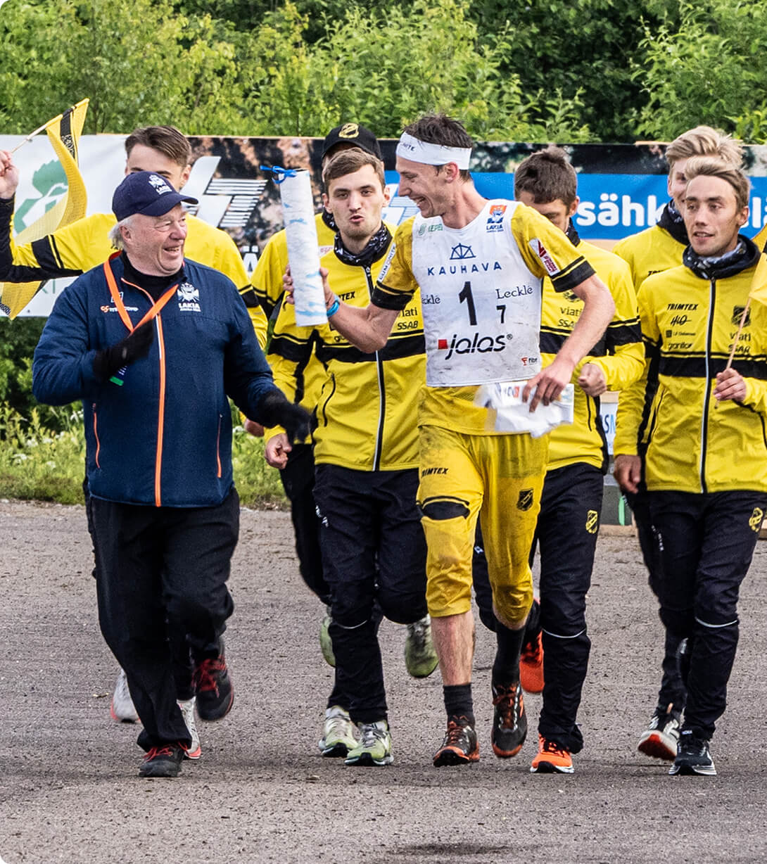 A group of male athletes in yellow and black uniforms, some smiling and running, celebrate with a coach in a blue jacket on a paved area with green trees in the background. One athlete holds a rolled-up paper.