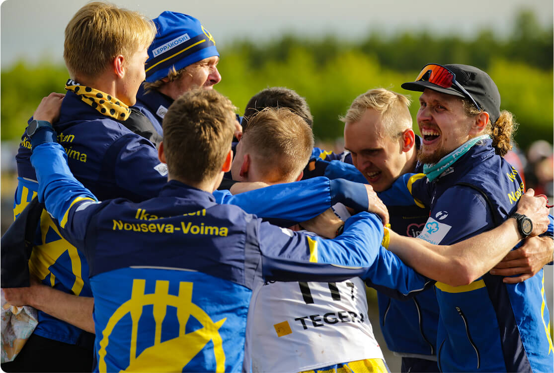 A group of six men wearing matching blue and yellow jackets huddle together outdoors, smiling and celebrating as they embrace each other. Green trees and blurred figures appear in the background.
