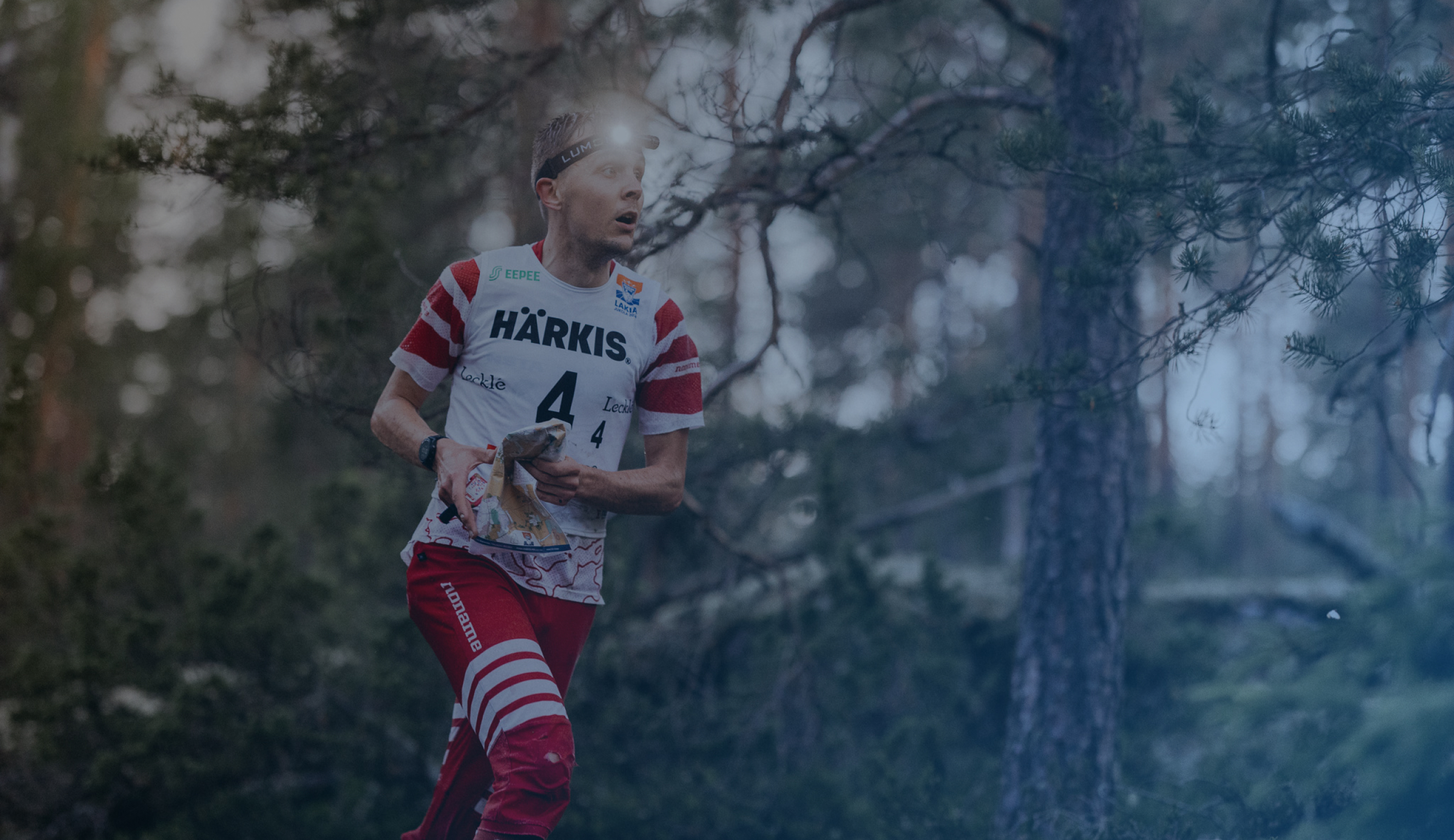 Otsalamppu ja punaiset raidalliset housut päässään oleva mies juoksee metsän läpi kartta ja kompassi kädessään suunnistuskilpailun aikana.