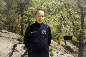 Valmentajan päivänä kiitetään koutsia. Nuori mies seisoo seinän edessä, jossa on maisemavalokuva avokalliosta ja männyistä. Miehellä on yllään Sotkamon lukion sininen pusero.