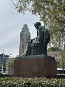 Sivukuvassa istuvan miehen patsas graniittijalustalla. Taustalla näkyy lehdessä olevan koivun oksia sekä korkea tornirakennus.