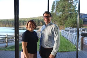 OP Kainuu on mukana Vuokatti-Jukolassa 2027 avainkumppanina. Nainen ja mies seisovat lasisen ikkunaseinän edessä. Ulkona näkyy lammen reuna, kaukana kohoava vaara ja ikkunan takana ulkoterassi.