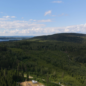 Droonilla otettu kesäinen maisemakuva. Kuvassa näkyy laaja havumetsäalue sekä taustalla kohoava vaara. Taivaanrajassa näkyy kaistale vesistöä. Taivaanrannassa on muutamia valkoisia pilviä. Kuvan alalaidassa näkyy pieni peltoalue, jolla on ihmisiä ja telttoja Esi-Jukolan kilpailukeskuksessa.
